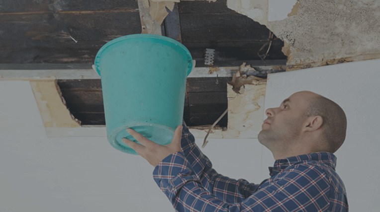 Man holding a bucket underneath a large leak from a very damaged ceiling