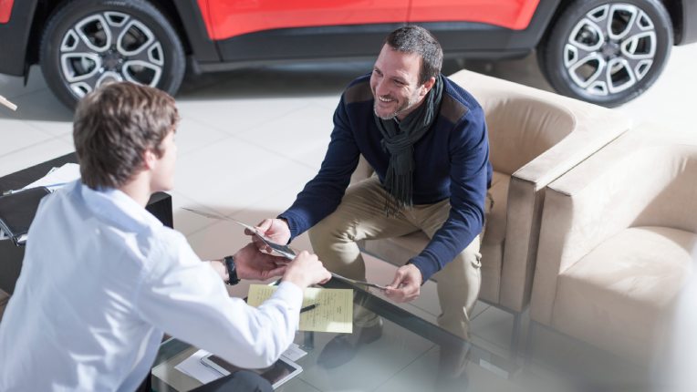 Two people sat opposite one another in a car sales room looking at a document with a red car in the background. Taken from a side angle from above.