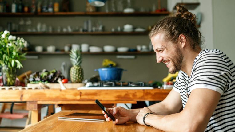 Caucasian person in a striped t-shirt sat at their kitchen table smiling at their smartphone whilst holding it